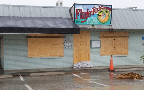 Boarded-up Flagler Fish Company restaurant with sandbags, hurricane-prepped storefront and empty parking lot under cloudy sky