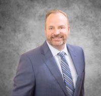 Smiling middle-aged bald man in blue suit and striped tie, professional corporate headshot against gray background