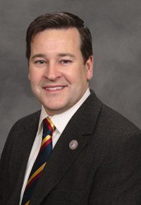 Professional headshot of a smiling man in a dark suit jacket and striped tie, positioned against a neutral gray background