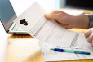 A hand holds a document near a laptop and blue pen on a wooden desk, focusing on reviewing or signing paperwork