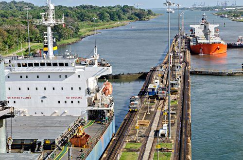 Large cargo ships passing through the Panama Canal's lock system featuring green shoreline and industrial infrastructure visible