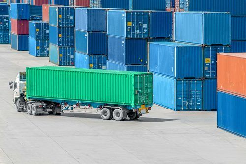 Green shipping container loaded on a trailer amid stacks of blue and red cargo containers at an industrial shipping yard