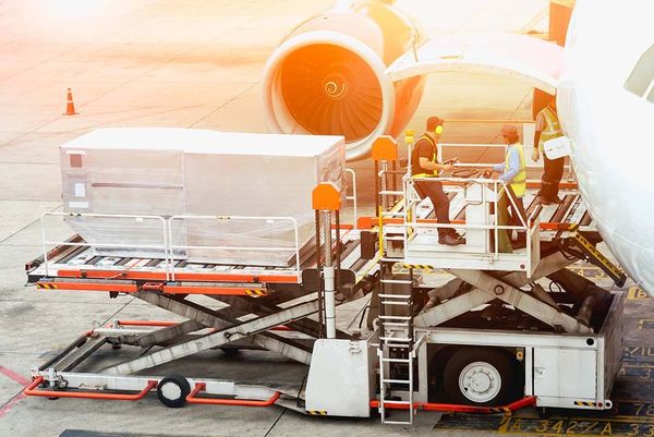 Ground crew loads cargo onto scissor lift beside orange-tipped aircraft engine on sunlit tarmac