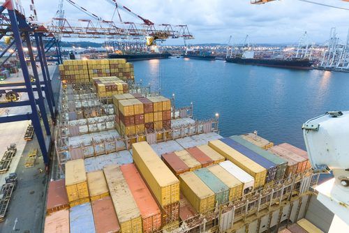 Stacked multicolored shipping containers on cargo ship deck with port cranes and blue water in background at dusk