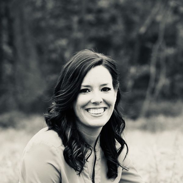 Smiling woman with wavy dark hair in black and white, standing in a blurry outdoor setting with trees in the background