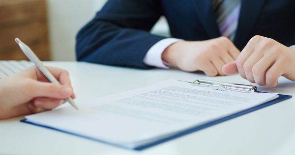 Two hands holding pens prepare to sign a document on a clipboard featuring one person wearing a navy blue suit jacket