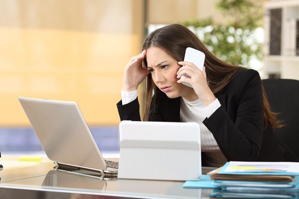 A stressed professional in a black blazer looks worried while working on a laptop and holding a white phone