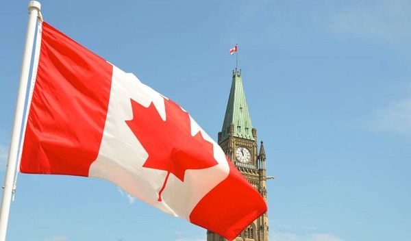 Canadian flag waves in foreground with Parliament's Peace Tower and clock visible against a clear blue sky in Ottawa