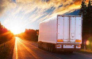 Sunset-lit semi-truck trailer driving down an empty highway with trees silhouetted against a golden, dramatic sky