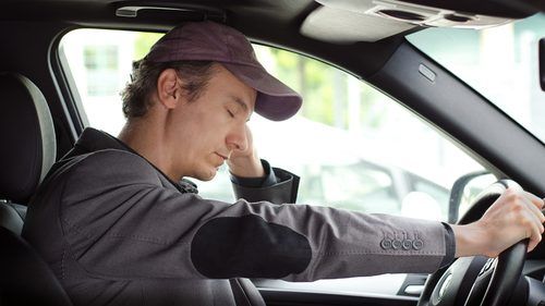 A tired-looking person in a dark cap and suit jacket slumped over the steering wheel, appearing exhausted while driving