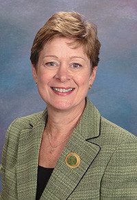 Professional headshot of a smiling woman in a green blazer with a blurred background, wearing a circular pin on her lapel