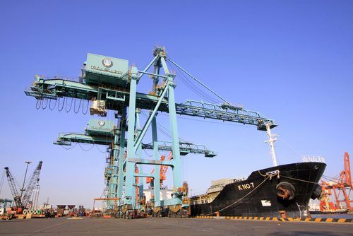 Large teal cargo crane looms over a black container ship in a bustling industrial port with orange cranes and clear blue sky