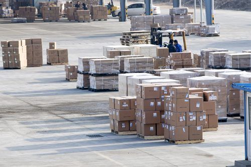 Stacked cardboard shipping boxes on pallets in a large warehouse loading dock with a forklift in the background