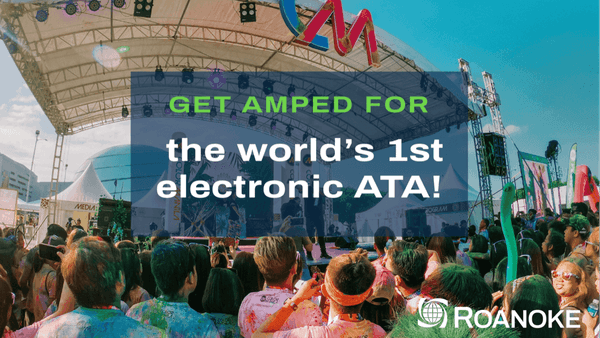 Colorful crowd gathered at an outdoor stage with "Get Amped for the world's 1st electronic ATA!" banner, vibrant