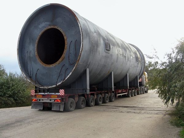 A massive industrial cylindrical tank transported on a multi-axle flatbed trailer along a tree-lined road