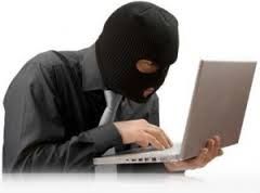 A masked individual in dark clothing using a laptop, suggesting cybercrime or hacking activity