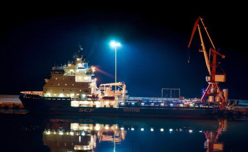 Illuminated icebreaker ship docked at night featuring bright blue light and red cargo crane reflecting in calm water