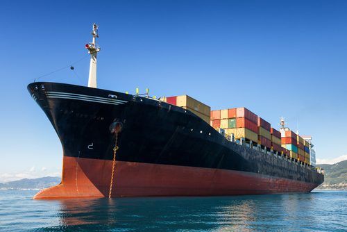 Massive cargo ship loaded with colorful shipping containers, anchored on blue water with mountainous coastline in background