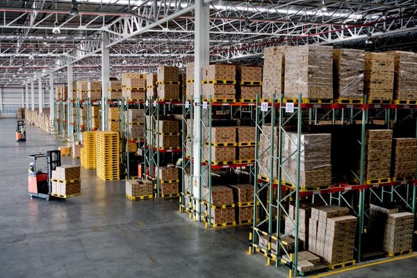 Large warehouse interior with tall shelving racks, stacked cardboard boxes on yellow pallets, and red forklifts