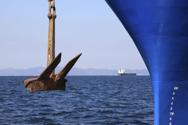 Rusted ship's anchor hanging from a chain featuring a blue ship's hull and distant cargo vessel on a calm blue ocean