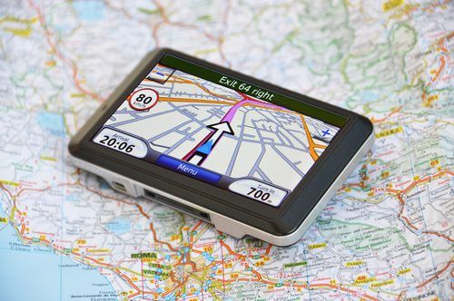 GPS navigation device displaying a digital map, sitting atop a physical paper road map with colorful geographical details