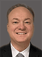 A professional headshot of a smiling middle-aged man in a dark suit and white shirt against a neutral gray background