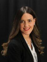 Smiling professional woman with long brown hair wearing a black blazer and white shirt against a dark background