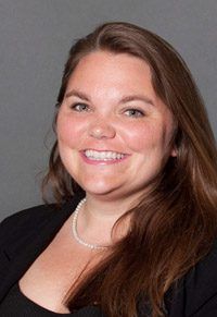 A smiling woman with long brown hair wearing a black top and pearl necklace against a gray background