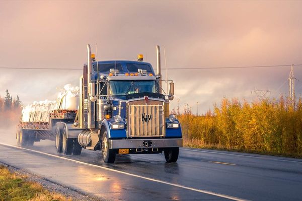 Blue Kenworth semi-truck hauling a flatbed trailer drives on a misty road at sunrise, surrounded by autumn-colored foliage