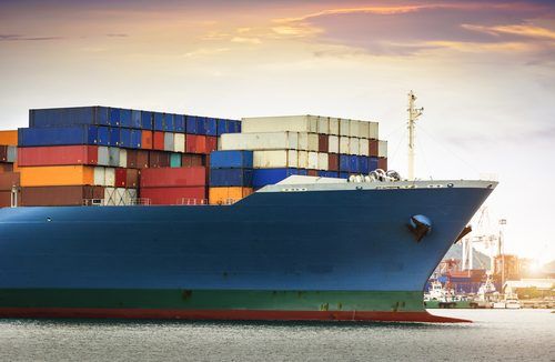 Massive container ship loaded with colorful shipping containers, sailing at sunset with a smaller vessel nearby