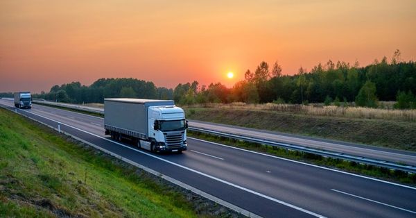White semi-truck driving on a highway at sunset featuring green forests and golden fields alongside the road