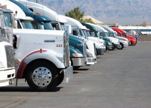 Row of parked semi-trucks in various colors lined up in a truck lot with mountains in the background