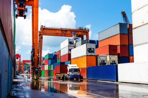 Colorful shipping containers stacked high featuring gantry cranes and a wet, reflective dock under a bright blue sky