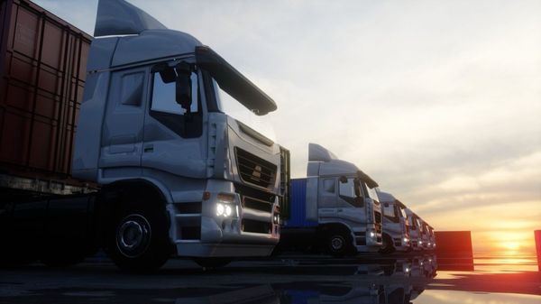 White semi trucks lined up at sunset featuring shipping containers and headlights illuminated against an orange sky