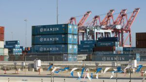 Stacked blue Hanjin shipping containers with red cargo cranes in background at industrial port or shipping terminal
