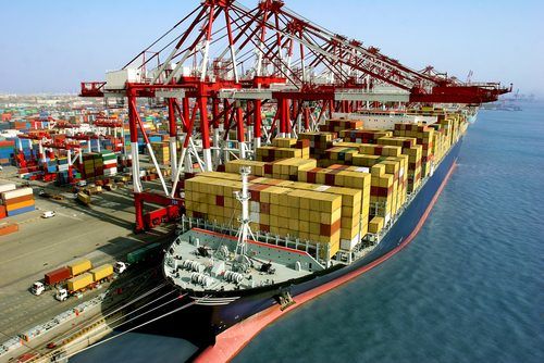 Massive container ship loaded with colorful shipping containers, docked at a busy port with large red gantry