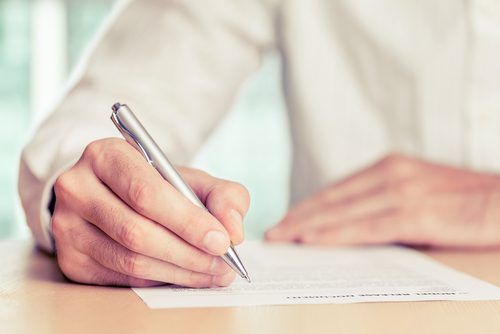 A hand holding a silver pen, writing on a white document with a blurred beige and white background