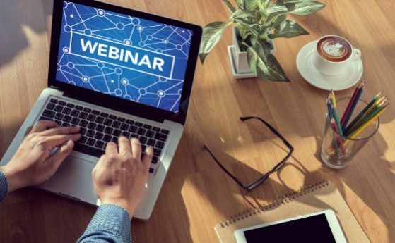 Laptop screen displays "WEBINAR" over network graphic featuring hands typing, coffee, plant, glasses, and notebook on wooden desk