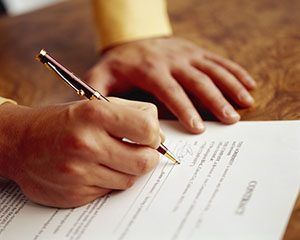 A hand holding a gold-tipped pen is shown writing on a document with printed text, resting on a wooden surface
