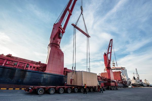 Large red cargo cranes lift a beige shipping container onto a multi-axle trailer at an industrial port under a blue sky