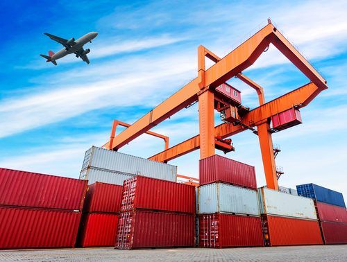 Orange gantry crane loading red and gray shipping containers with airplane flying overhead against bright blue sky