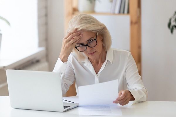 An older professional with blonde hair and glasses looks stressed while reading a document at a white laptop