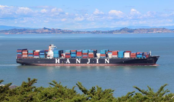 Hanjin cargo ship loaded with colorful shipping containers sailing on calm blue waters with mountainous coastline