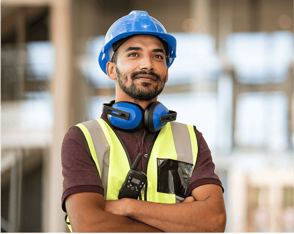 Smiling construction worker with blue hard hat, safety vest, and noise-canceling headphones around neck