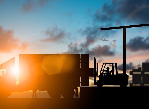 Silhouetted forklift loading shipping containers at sunset featuring dramatic orange and blue sky in background