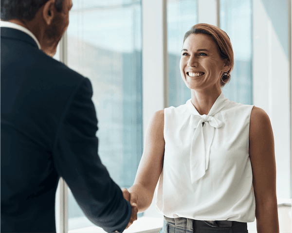 Smiling professional in white sleeveless blouse shaking hands with colleague in dark suit, bright office background