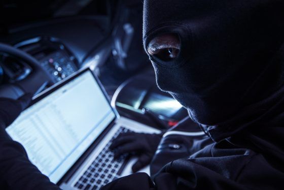 Masked figure in dark hoodie using laptop, dramatically lit by screen's glow, suggesting cybercrime or hacking activity
