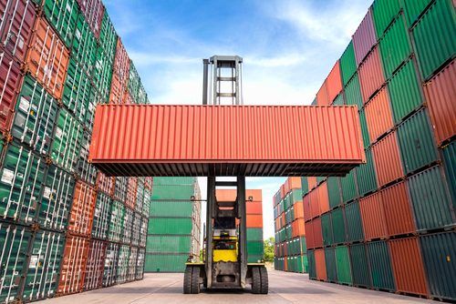 Yellow forklift moves red shipping container amid green and orange cargo containers at industrial port
