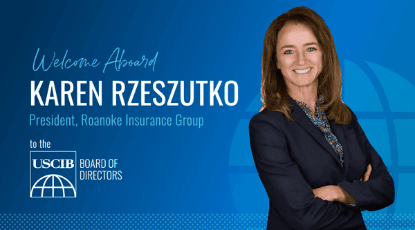Smiling businesswoman in navy suit stands confidently against blue gradient background welcoming her to USCIB