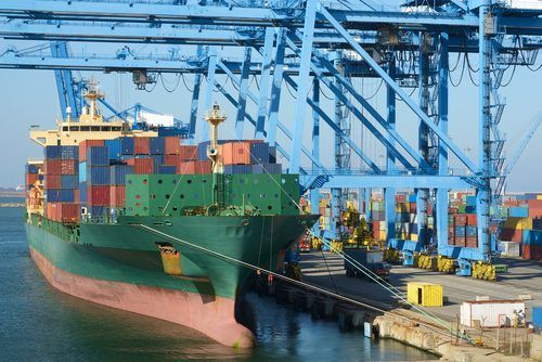 A massive green and pink cargo ship loaded with colorful containers docked at an industrial port with blue cranes